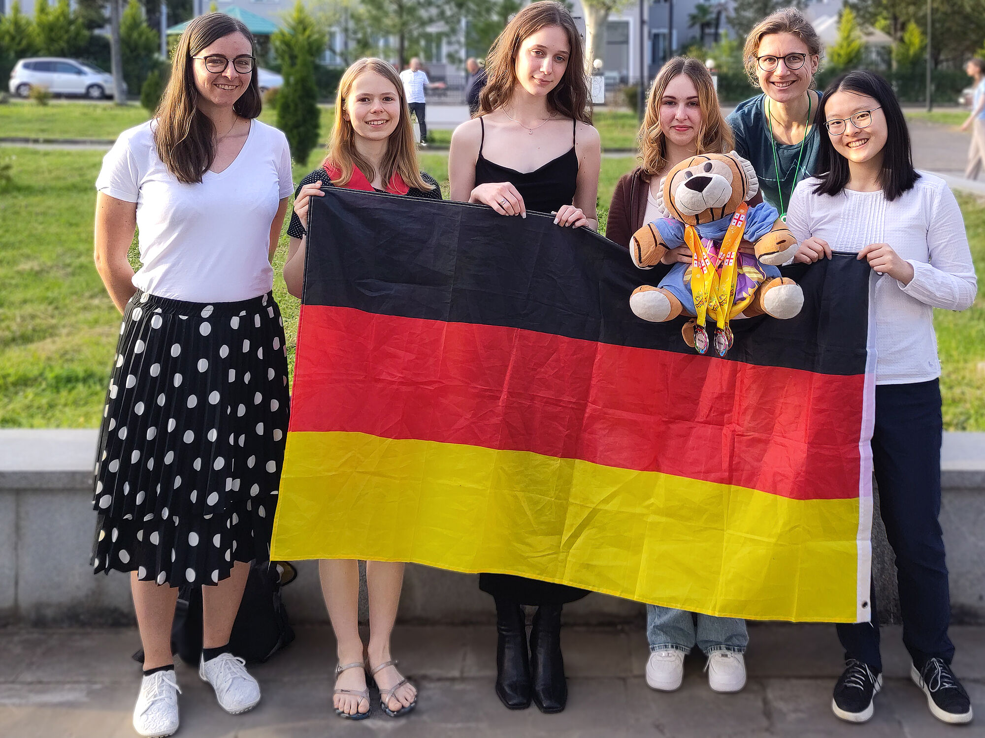 Die vier Teilnehmerinnen an der EGMO 2024 zusammen mit den beiden Delegationsleiterinnen, der deutschen Flaggs und dem Maskottchen "MathemaTigerin", der mehrere Medaillen umgehängt wurden. 